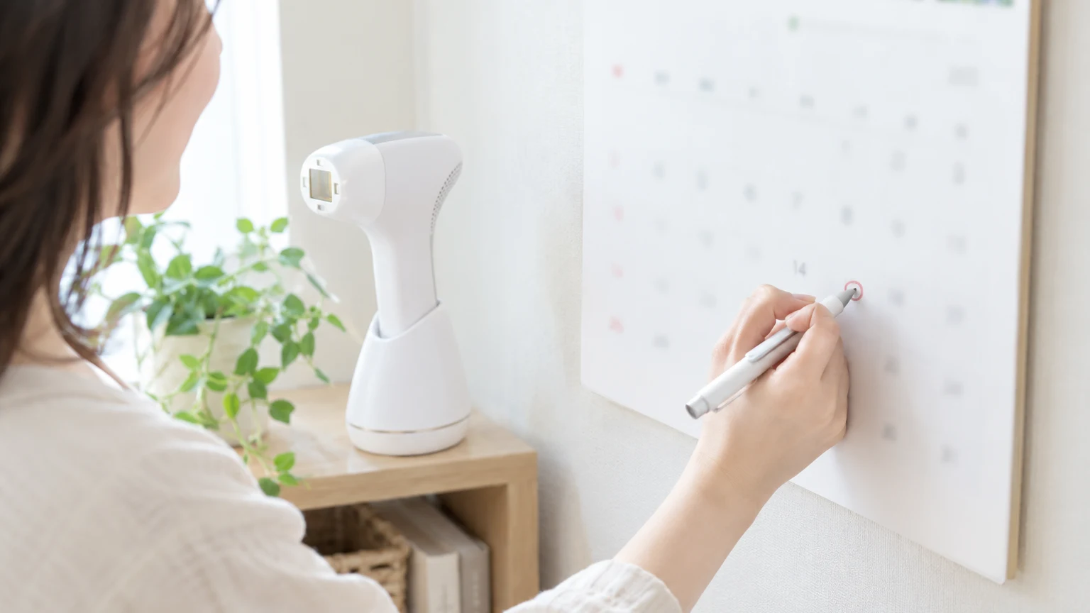 カレンダーにペンで丸をつけている手元、そばに白い光美容器が置かれている
