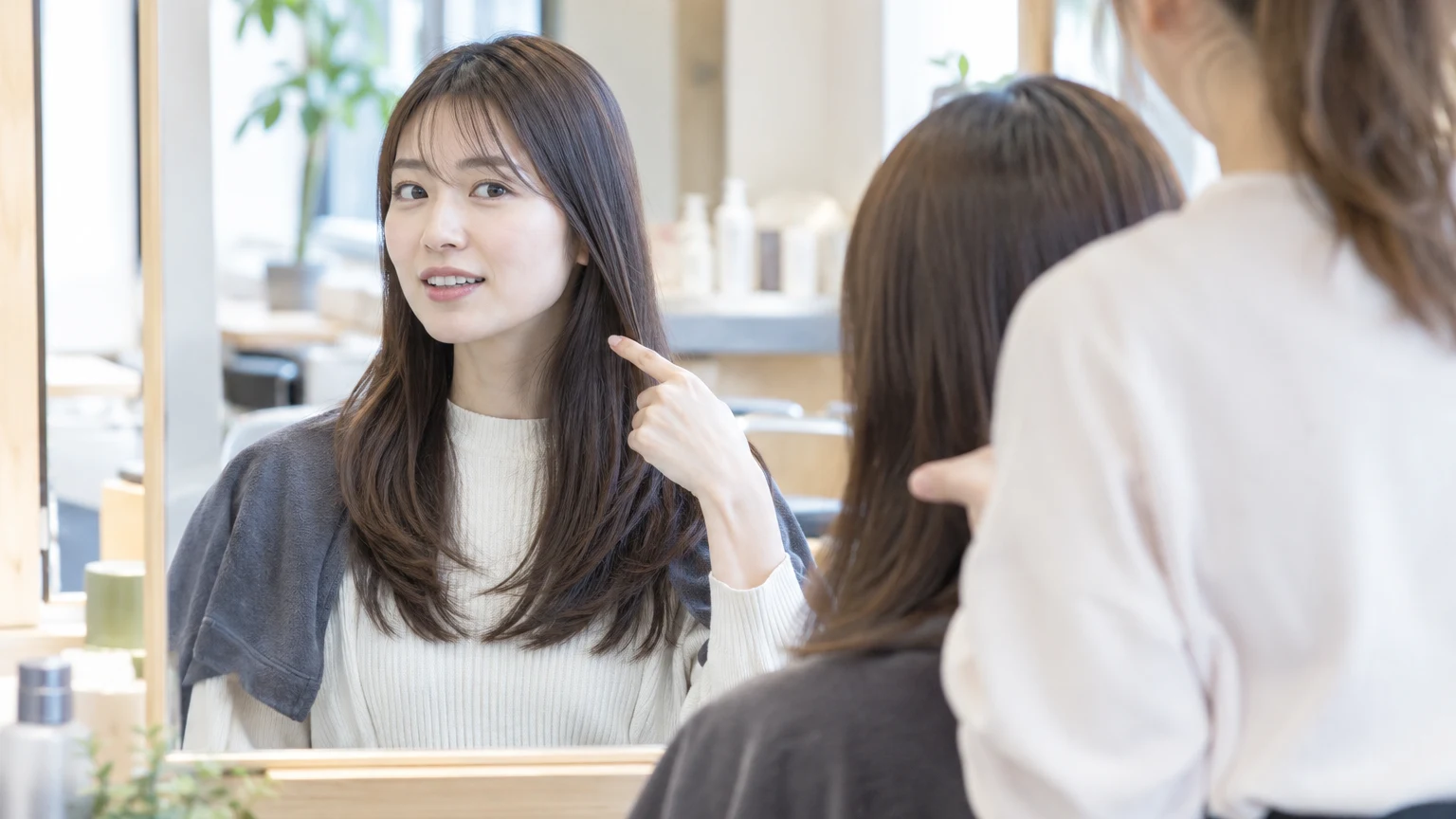 美容室の椅子で、乾いた髪のまま鏡越しに美容師へ髪の高さを示しているところ