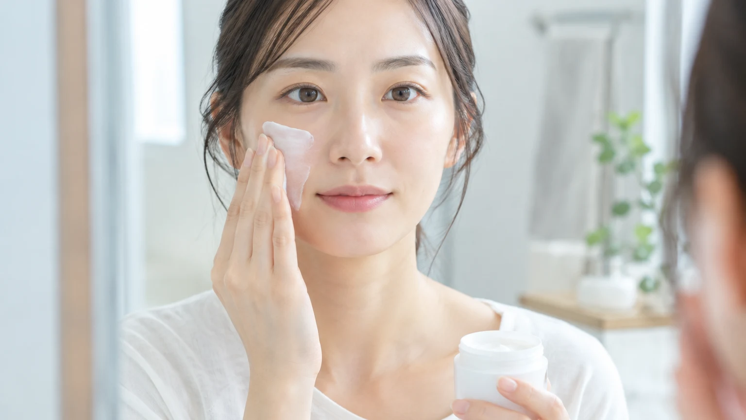 洗面所の鏡の前で、片手にクリームの容器を持ち、指先で頬にクリームをのばしている女性