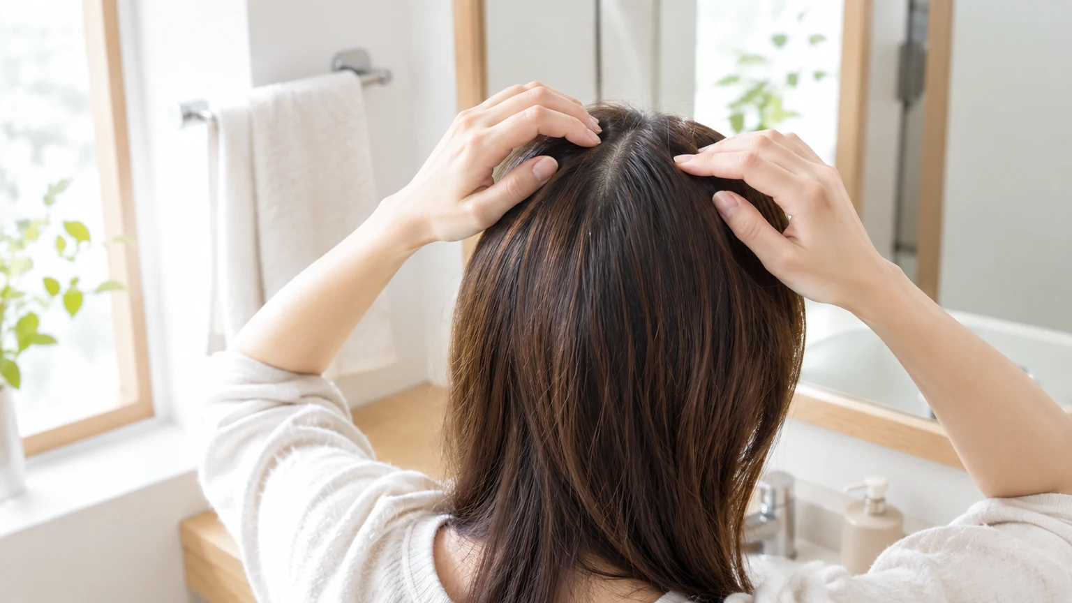 朝の洗面所の鏡の前で、分け目を指で少し開いて根元の色を確かめている手元