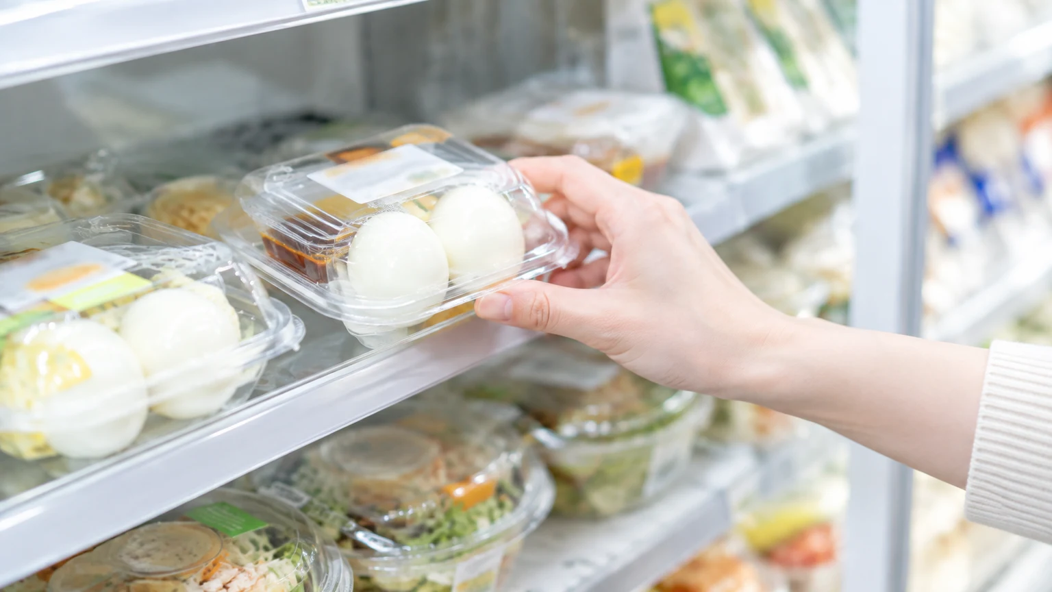 コンビニの冷蔵の棚で、殻をむいてある味付きのゆで卵のパックを手に取っている手元