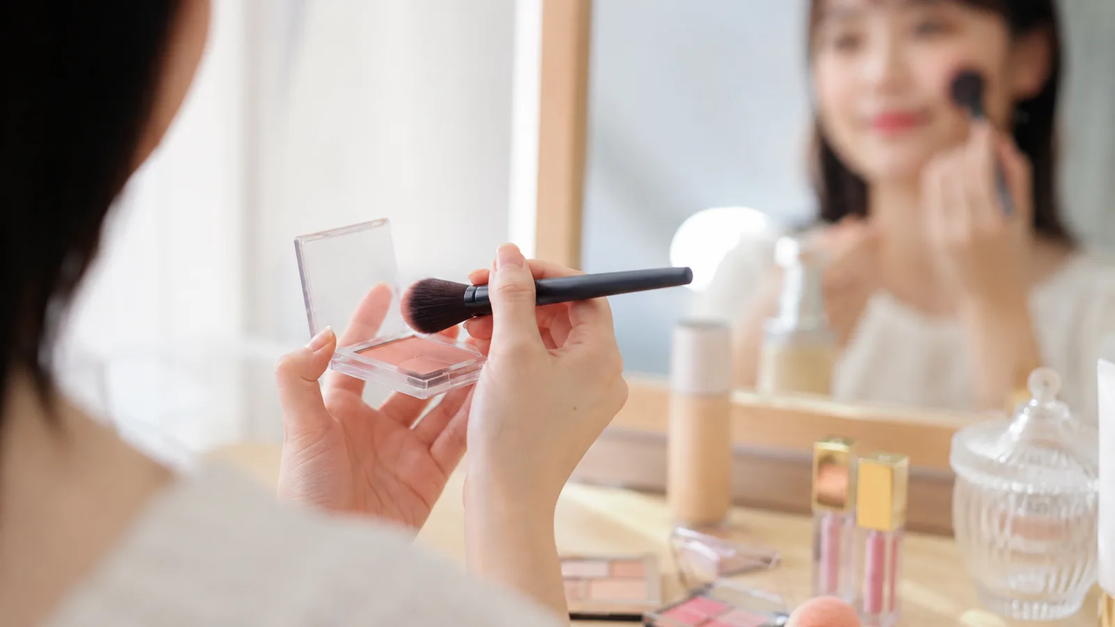 鏡の前でチークをブラシに取りメイクの色味を確認しながら仕上げていく女性