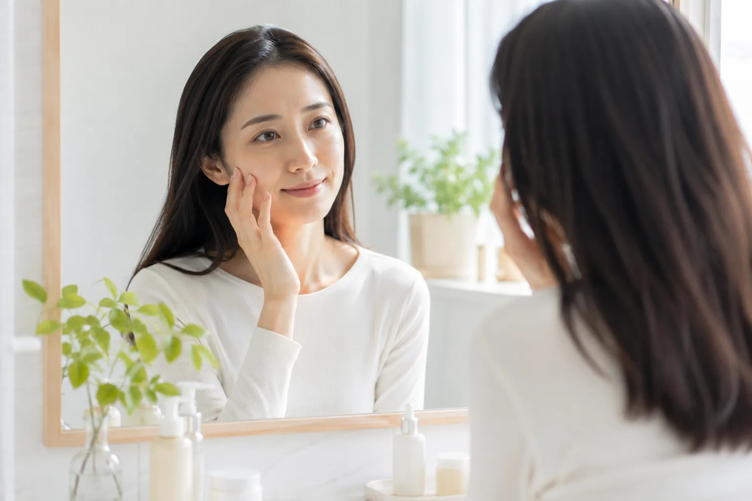 洗面所の鏡の前で頬に指先を添え、気負わず化粧水をなじませるように微笑む女性
