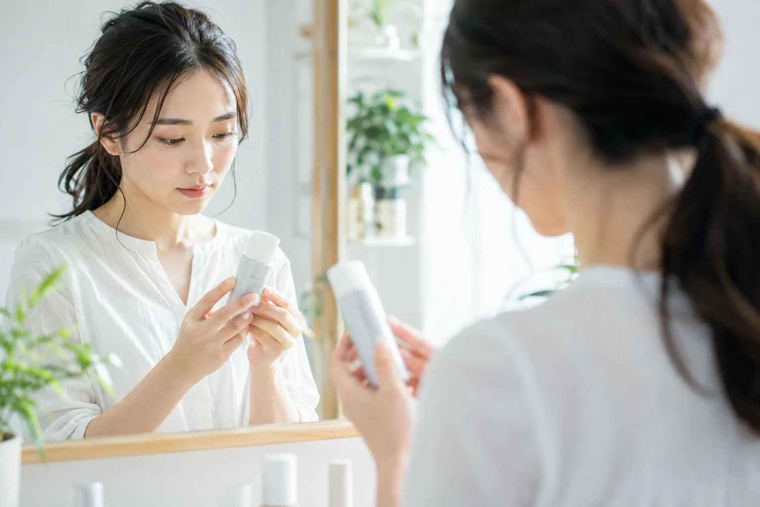 洗面所の鏡の前で、白い容器を両手で持ち、表示を確かめている女性の後ろ姿と鏡に映る姿