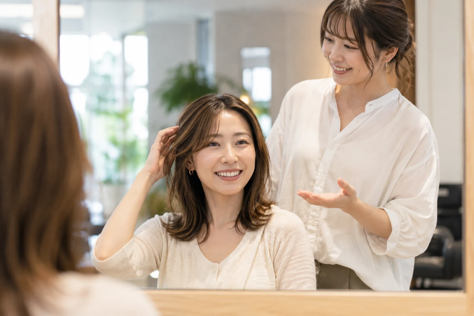美容室の鏡の前で、髪に手を添えて笑顔で話す客と、手のひらを向けて説明する美容師