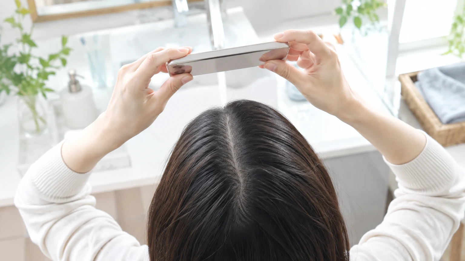 洗面所で、スマートフォンを頭の上にかざし、分け目を真上から撮ろうとしている頭頂部と手元