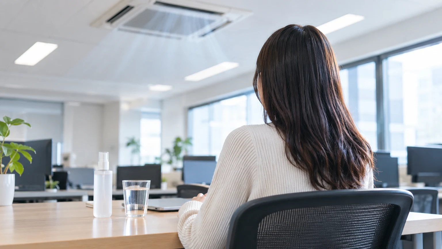 エアコンの吹き出し口の下のオフィスの席に座る女性の後ろ姿と、机の上のミストボトルと水の入ったコップ