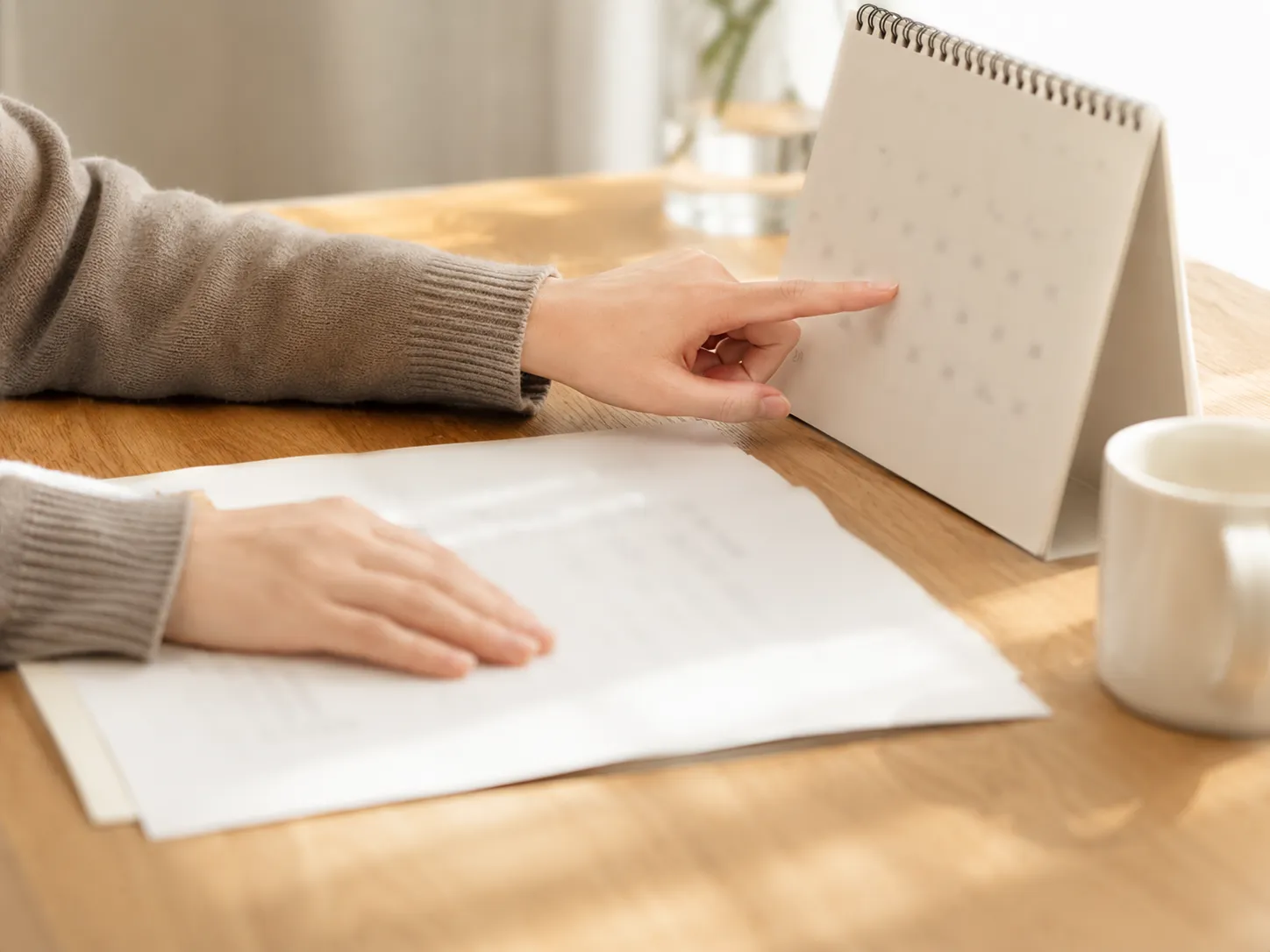 木のテーブルで卓上カレンダーの日付を指でなぞり、書類と見比べている手元