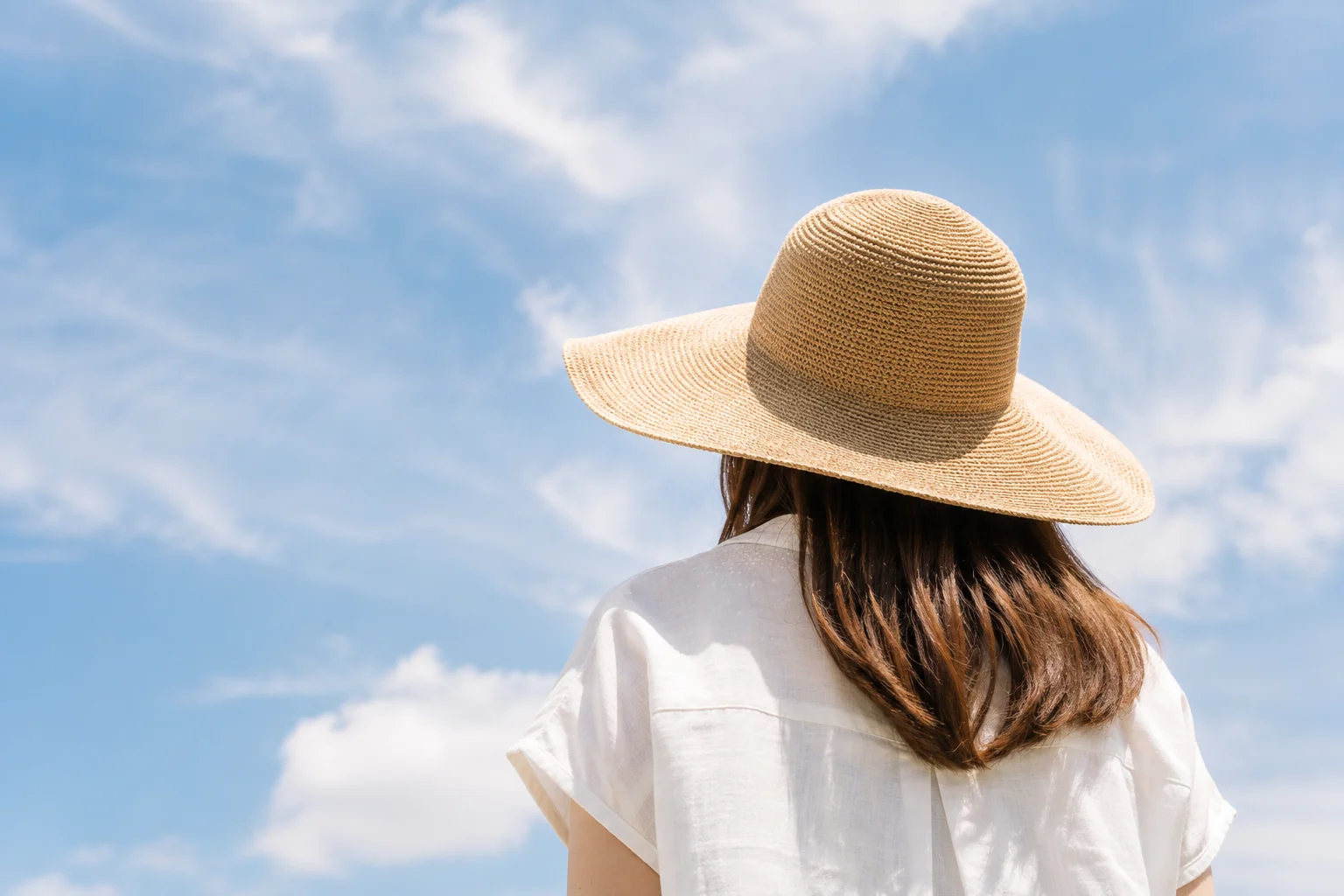 青空の下で、つばの広い麦わら帽子をかぶった肩までの髪の女性の後ろ姿